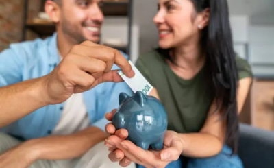 Pareja joven sonriente ahorrando juntos, colocando un billete de 100 € en una hucha, simbolizando la organización y planificación de las finanzas en pareja